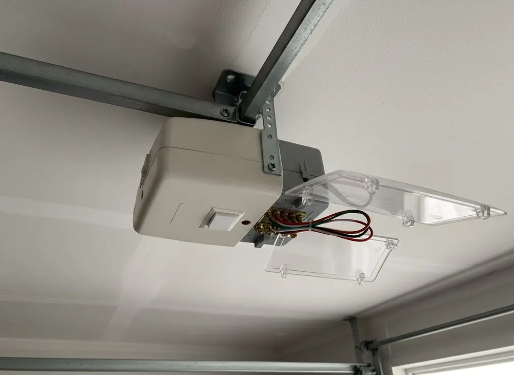 Opener motor head on a garage ceiling with its rear cover open showing the program button